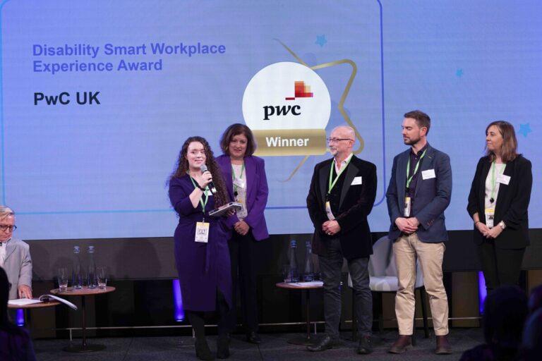 Group of 5 people on stage during a Disability Smart Awards ceremony. One is holding a trophy and speaking into a microphone.