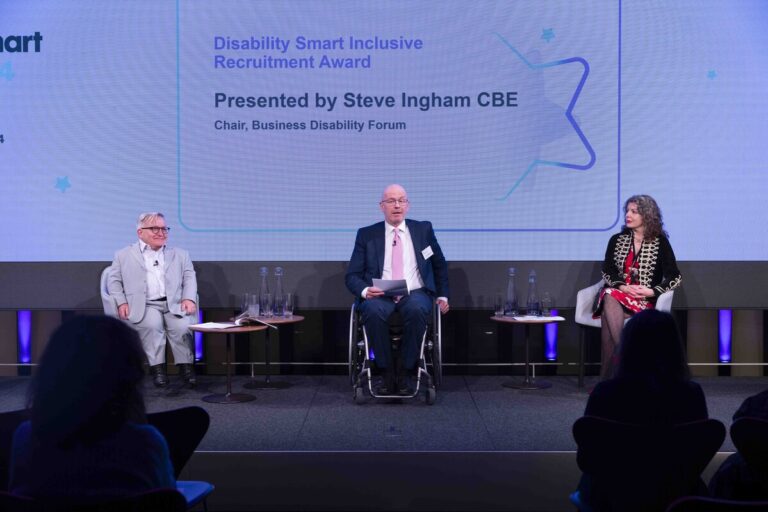 3 speakers, including a person of small stature and a wheelchair user, on stage during an event. Behind them is a screen with the Disability Smart Awards logo projected on it.