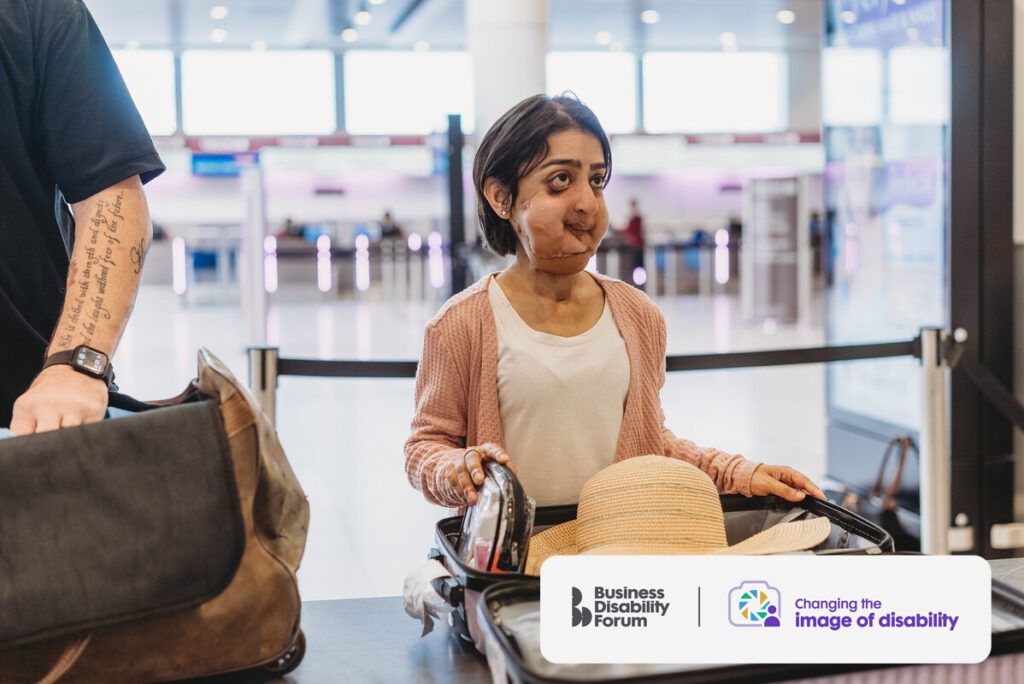 Person with a facial difference who is short in stature getting their baggage checked at the airport. Their suitcase is open in front of them and they're holding a toiletry bag.