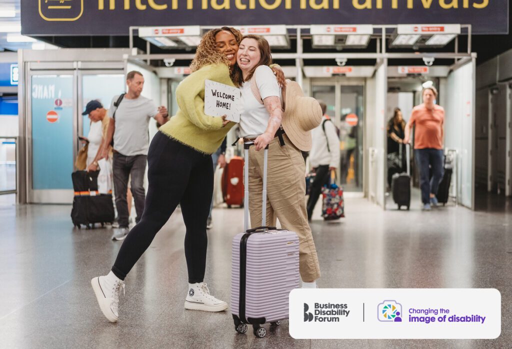 Person with scarring and a limb difference at the airport, being welcomed by a friend with a less-visible disability. The friend embraces them while holding a sign that reads 'Welcome home'.