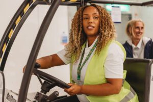 A woman with a less-visible disability (chronic illness) drives a passenger assistance vehicle at the airport. She is wearing a sunflower lanyard and a high vis vest. 