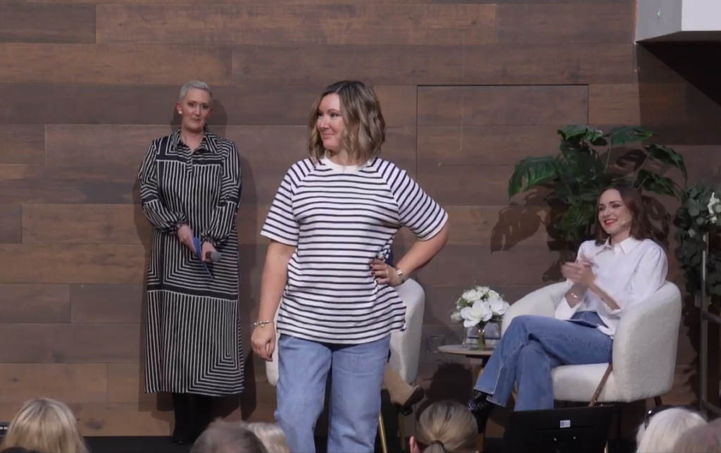 A white woman with blonde hair modelling clothes on a catwalk. She is wearing denim jeans and a stripped top and poses with a hand on her hip. Looking on from behind her are a woman holding a microphone and a woman sitting in an armchair, who's clapping.