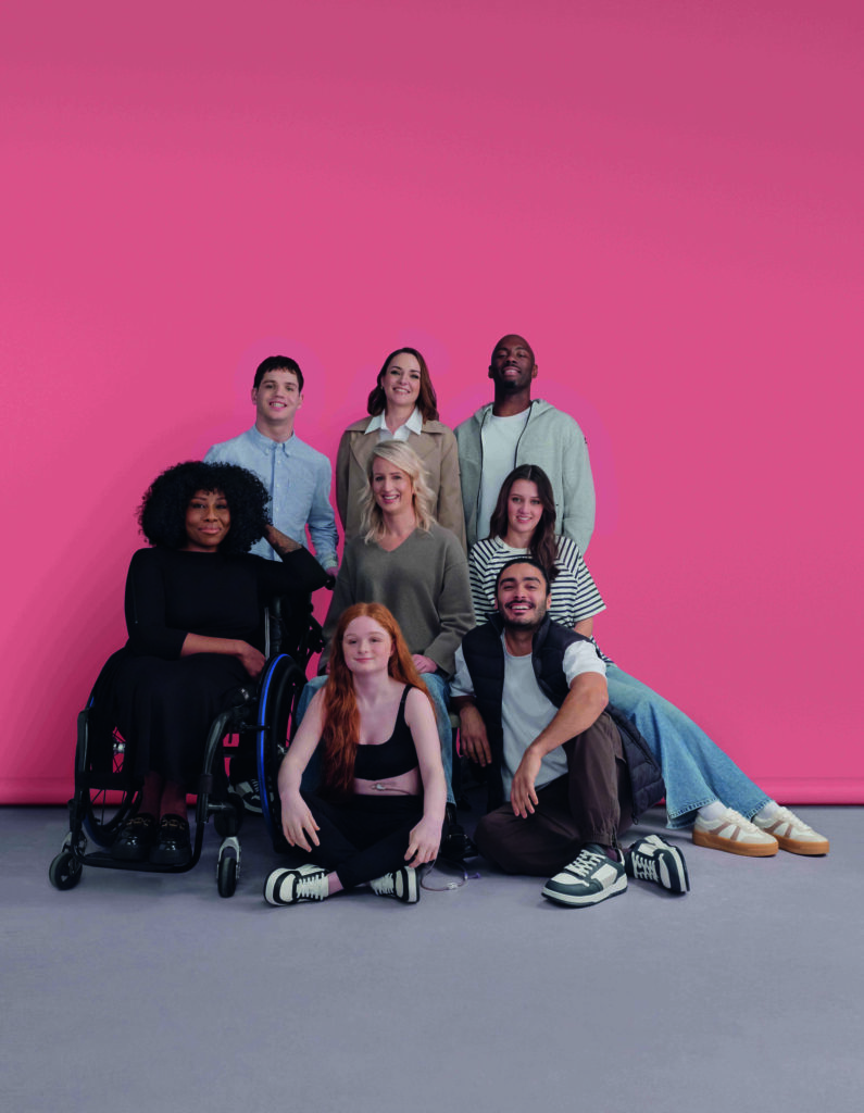 Group of 8 people with a range of disabilities posing against a pink background for a fashion shoot.