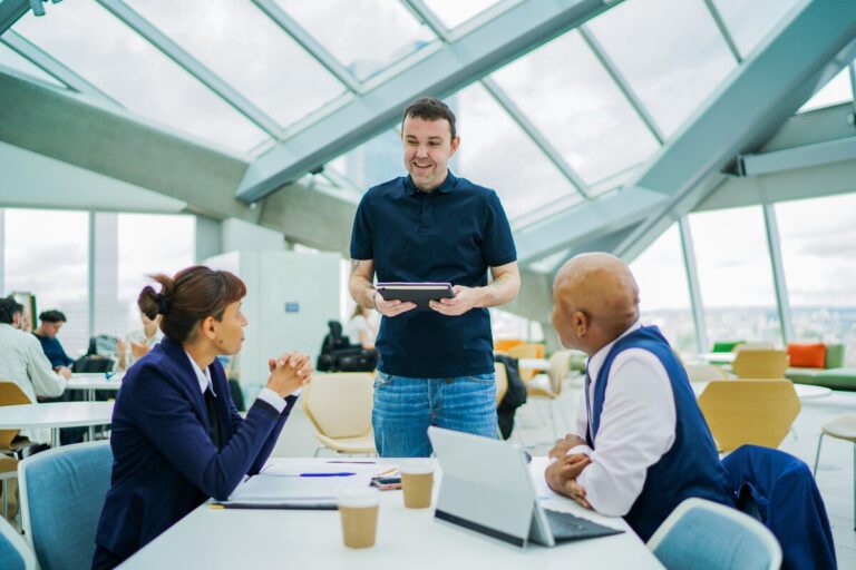 3 colleagues with less-visible disabilities talking. One professional is wearing a sunflower lanyard and standing while holding a notebook. The other two sit looking up at him.