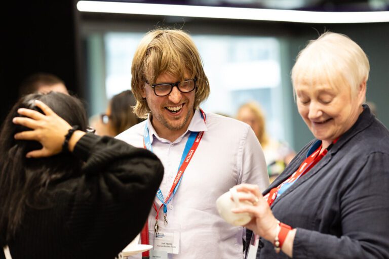 Man with a sight impairment who wears glasses smiles while talking to a delegate at an event. His support workers stands next to him, smiling as she checks her watch.