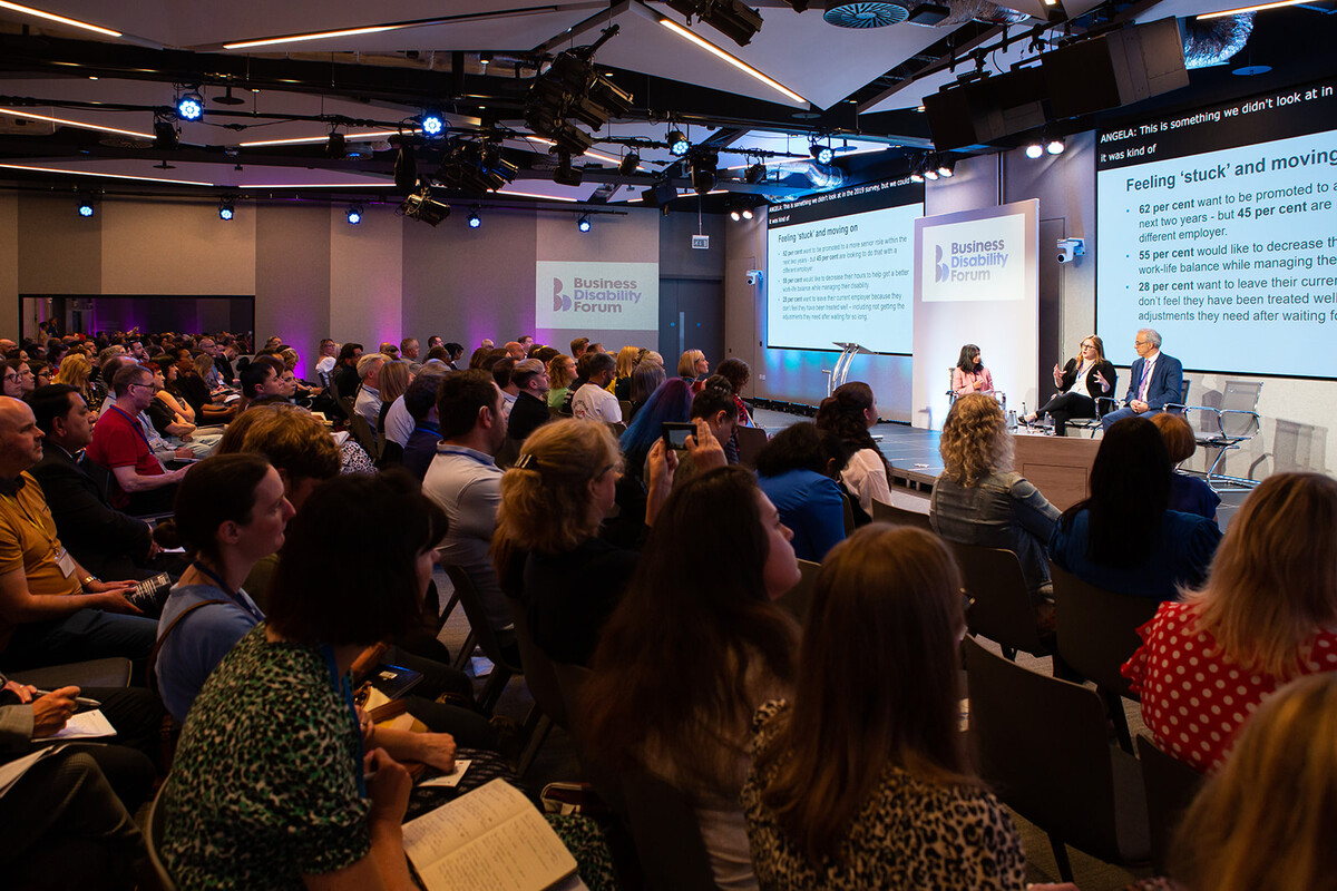 An audience listening to a panel of 3 people who are on stage at a conference.