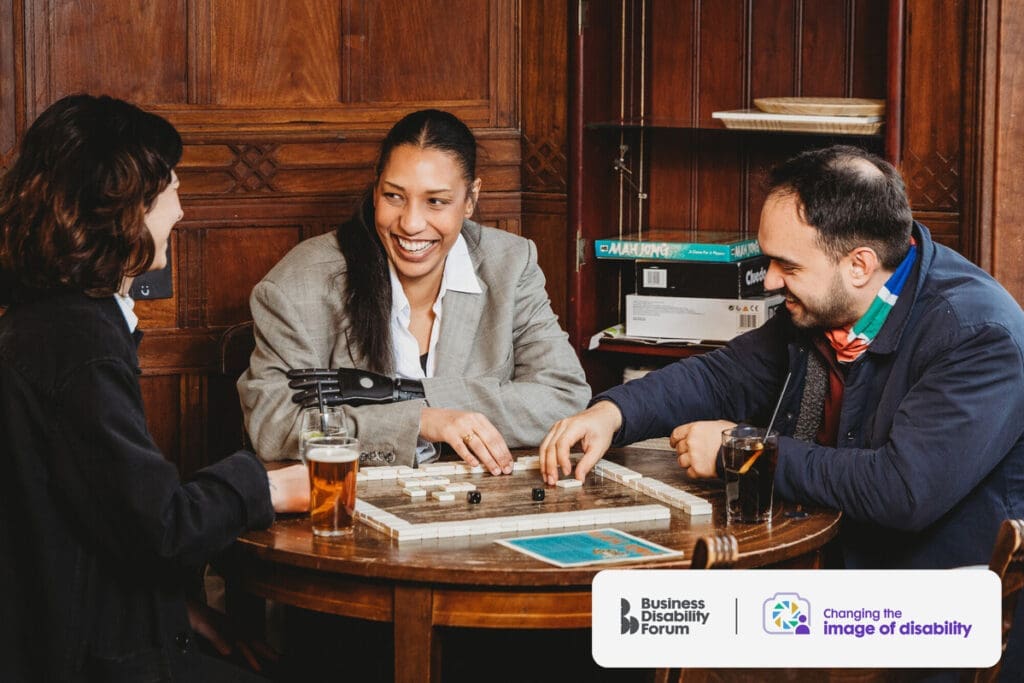 Friends with multiple disabilities play a board game in a pub. The woman in the centre wears a prosthetic arm and the man on the right has less-visible disabilities.